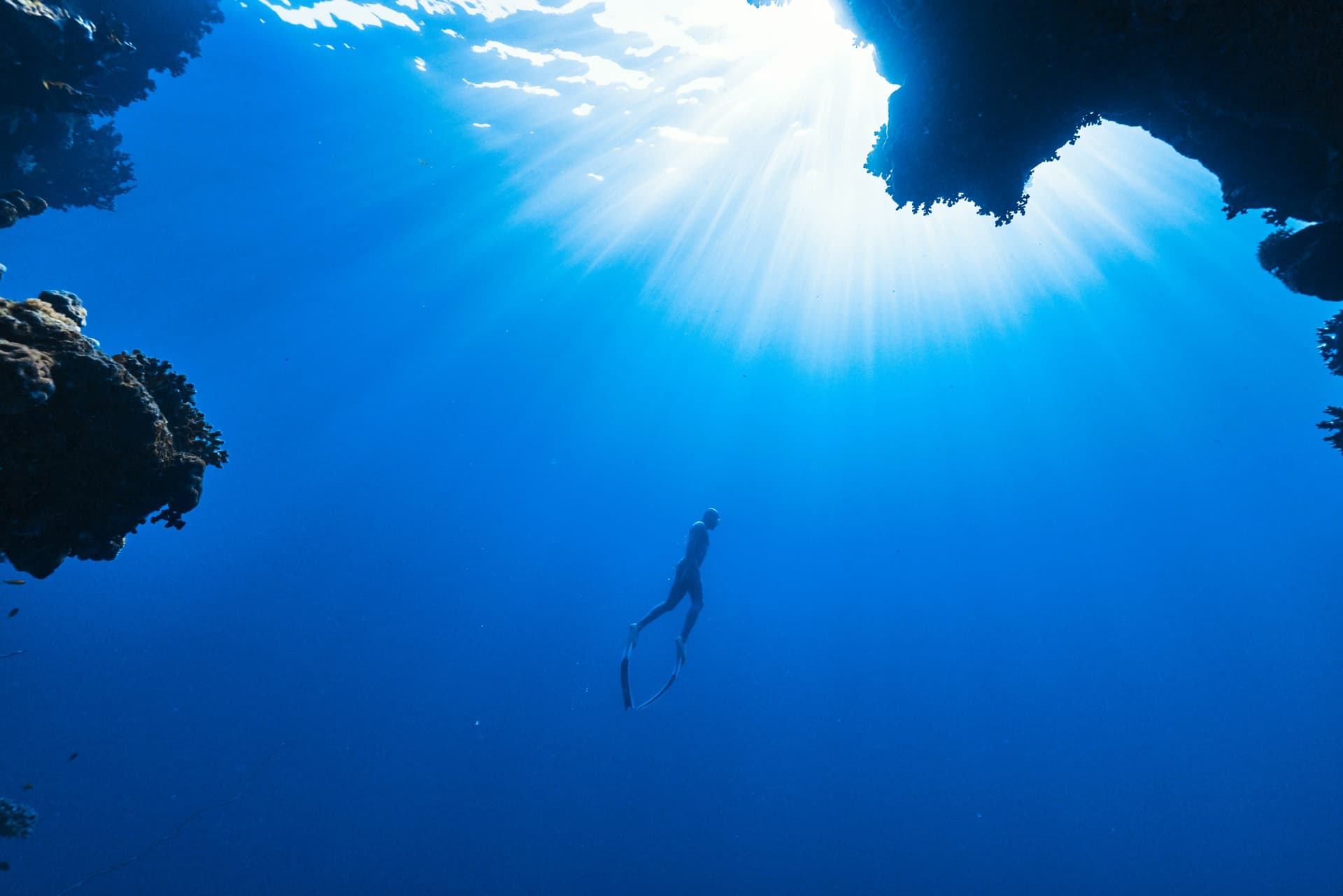 A freediver rising toward a sunburst through an opening in the reef